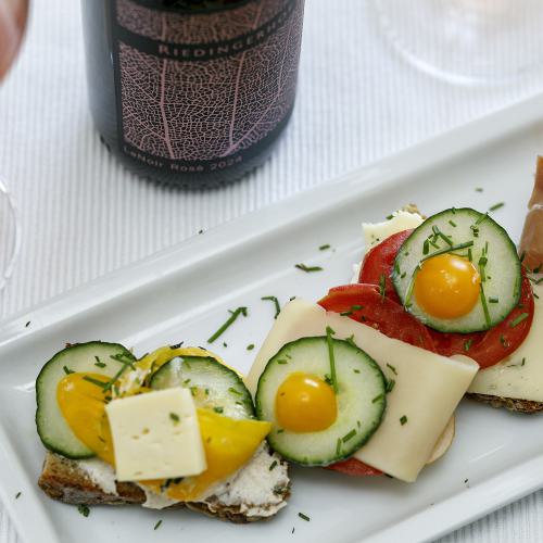 Liebevoll angerichtete Brotzeitplatte mit belegten Broten, Käse, Gurken, Tomaten und Südtiroler Speck, serviert mit einem Glas Rosé &ndash; dem hauseigenen LaNoir Rosé 2024 des Weinguts Riedingerhof in Meran.