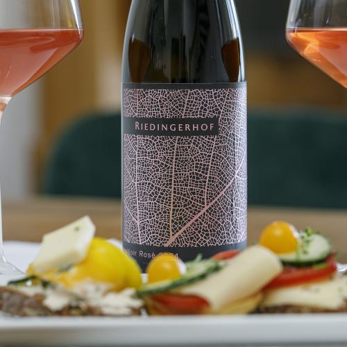 Liebevoll angerichtete Brotzeitplatte mit belegten Broten, Käse, Gurken, Tomaten und Südtiroler Speck, serviert mit einem Glas Rosé &ndash; dem hauseigenen LaNoir Rosé 2024 des Weinguts Riedingerhof in Meran.