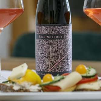 Liebevoll angerichtete Brotzeitplatte mit belegten Broten, Käse, Gurken, Tomaten und Südtiroler Speck, serviert mit einem Glas Rosé &ndash; dem hauseigenen LaNoir Rosé 2024 des Weinguts Riedingerhof in Meran.